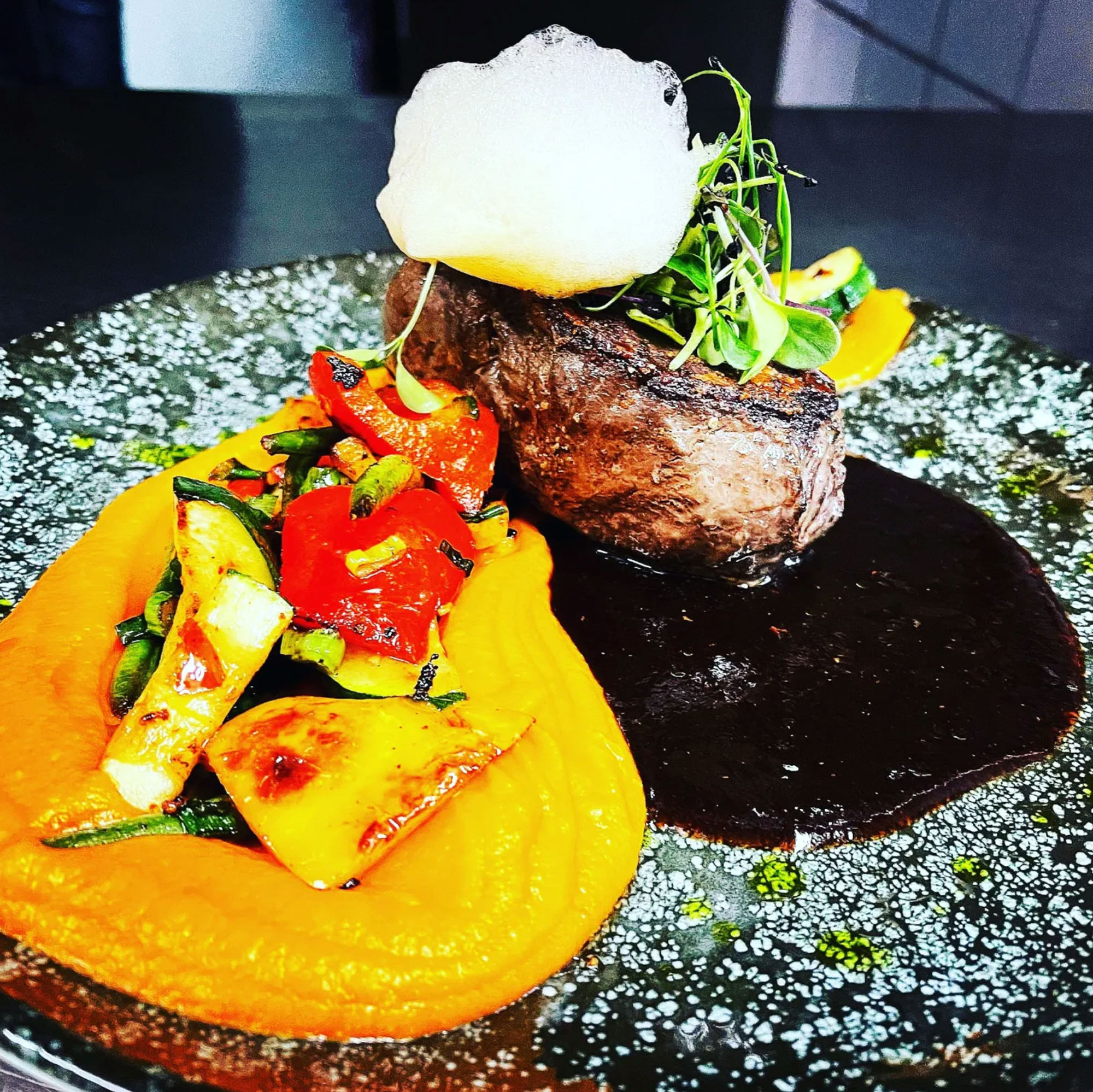 Rindermedaillon mit dunkler Jus, Kürbispüree und gegrilltem Gemüse, angerichtet auf dunklem Steinteller.