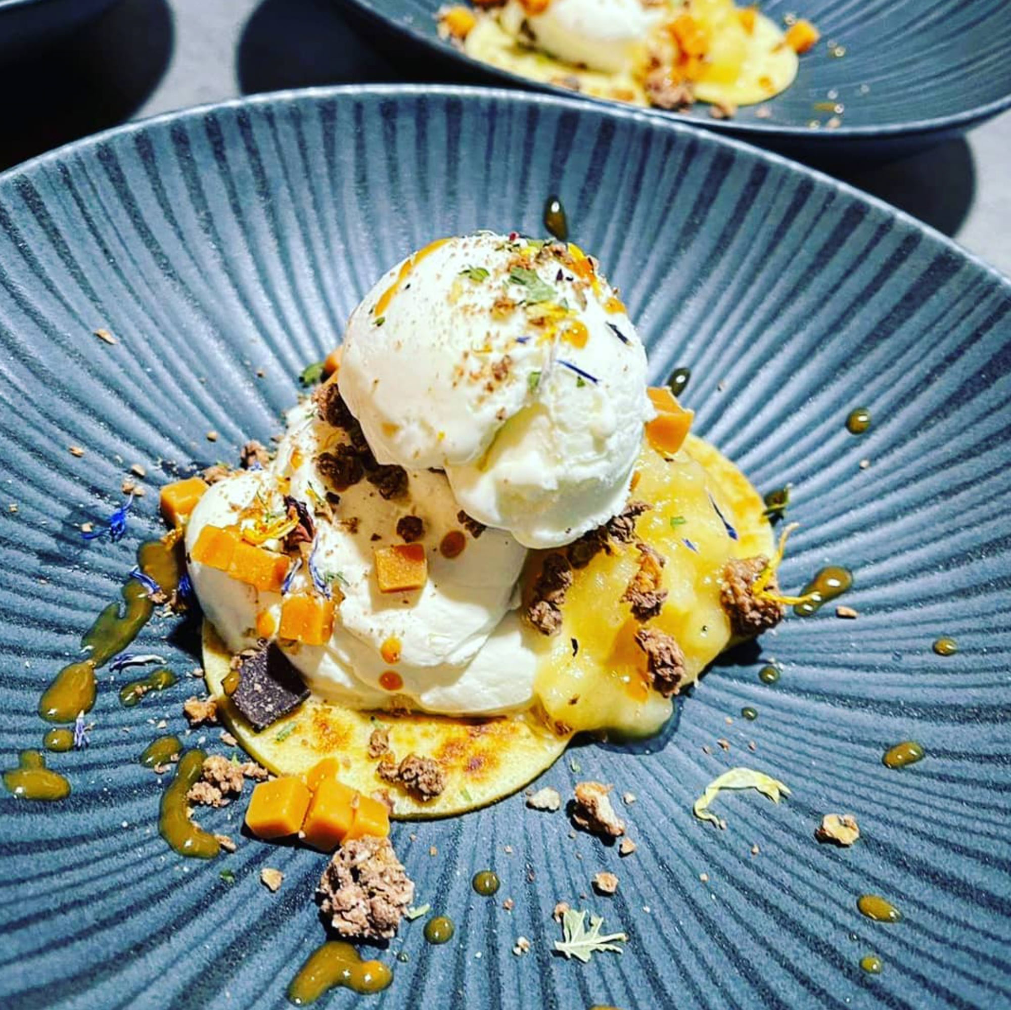 Dessert mit drei Eiskugeln, Karamellwürfeln, Streuseln und Fruchtspiegel auf blau geriffeltem Teller.