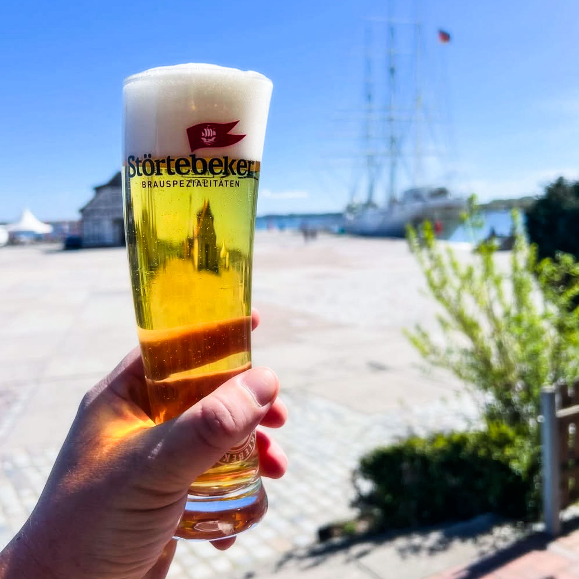 Hand hält ein frisch gezapftes Störtebeker Pilsener vor dem Stralsunder Hafen, dahinter die Masten eines Großseglers.